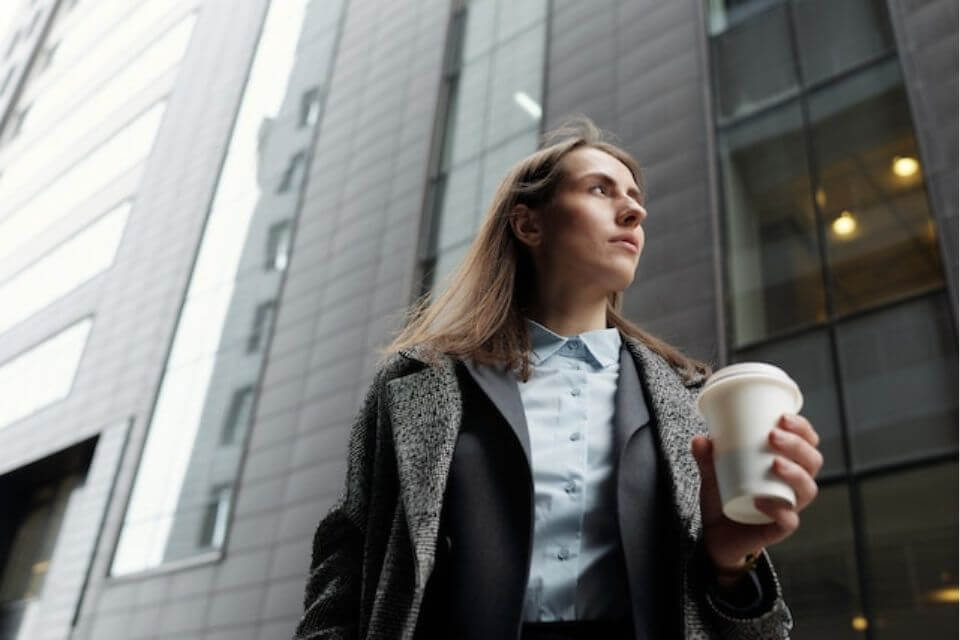 Photo shows a businesswoman walking with coffee against the backdrop of a high-rise building

